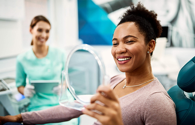 Happy female patient admiring her smile in mirror at modern dental office with dentist in background.